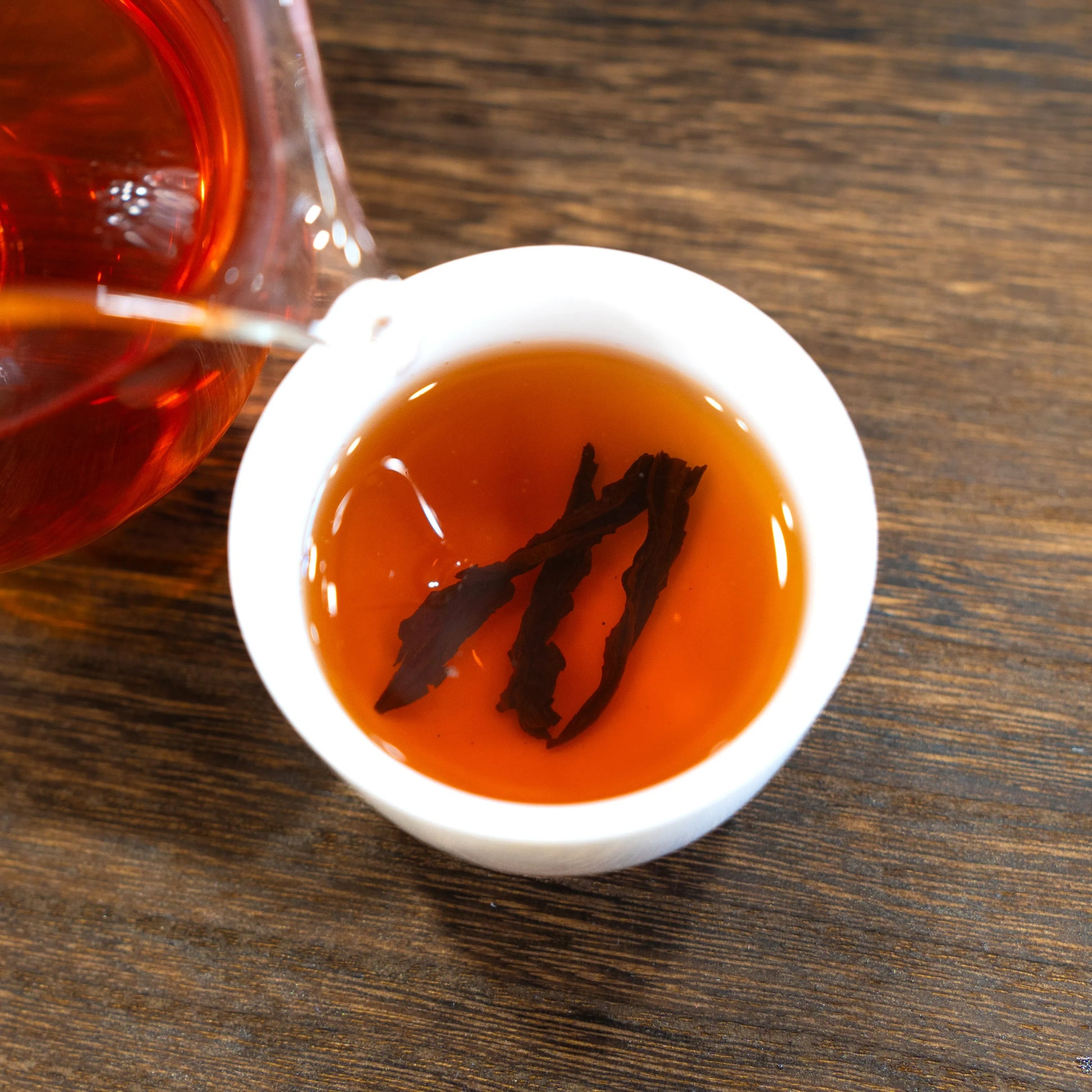 Rooteas iron arhat rock tea being poured into a small white cup with tea leaves on a wooden surface